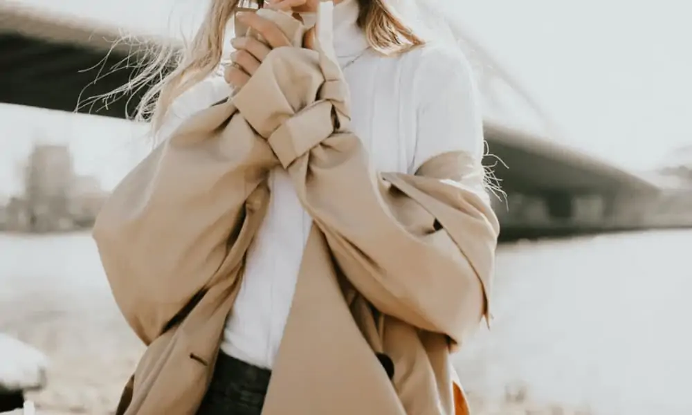 Woman wearing a white turtleneck cable knit sweater, with a trench coat, and black jeans. A good example of what to shop for when creating a sustainable and minimalist wardrobe.