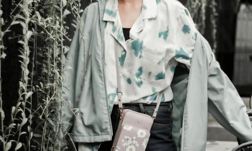 Woman wearing a printed button up top and green jacket with jeans and a small purse. 
